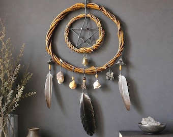May include: A wall hanging featuring two concentric woven willow rings, with a central dark pentagram. Suspended from the rings are a small brass bell, various natural stones, and several grey and brown feathers attached by dark cord.