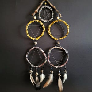 May include: A wall hanging featuring a central wooden branch supporting five circular hoops arranged in a vertical pattern. The hoops are wrapped in yellow and grey fibers, each containing a geometric string web. Natural feathers hang from the bottom.