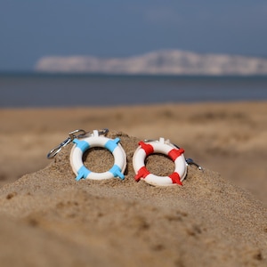 Könnte beinhalten: Zwei Miniatur-Rettungsring-Schlüsselanhänger, einer mit blauen Akzenten und der andere mit roten, liegen auf Sand. Die Schlüsselanhänger sind weiß mit farbigen Streifen, vor einem verschwommenen Strand- und Meereshintergrund.