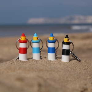 Puede incluir: Cuatro llaveros de faro de colores con rayas rojas, azules y negras, y tapas amarillas y negras, colocados en una playa arenosa. Cada llavero tiene un anillo plateado y una llave pequeña. El océano y el cielo están al fondo.