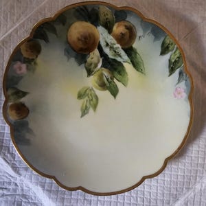 May include: A decorative, scalloped-edge plate with a gold rim. The plate features a hand-painted design of peaches, green leaves, and small pink flowers. The background is a soft yellow and blue.