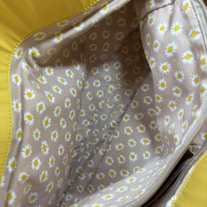 May include: A bright yellow tote bag with a daisy-patterned interior. The interior fabric is tan with a repeating pattern of white daisies with yellow centers. The bag has a zipper closure and a pocket.