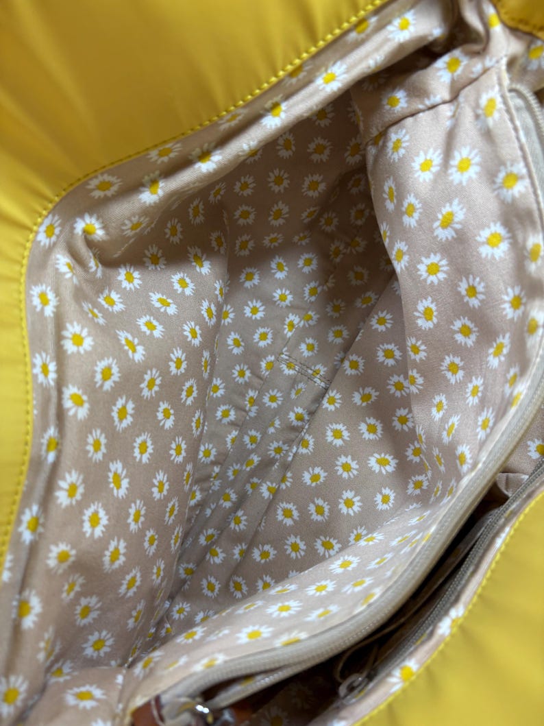 May include: A bright yellow tote bag with a daisy-patterned interior. The interior fabric is tan with a repeating pattern of white daisies with yellow centers. The bag has a zipper closure and a pocket.