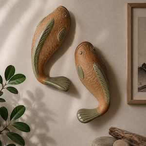 May include: Two wall-mounted fish sculptures crafted from textured tan ceramic with smooth, sage green glazed accents on the fins, gills, and tails. The fish are positioned vertically, each featuring a curved body shape and detailed scale patterns.