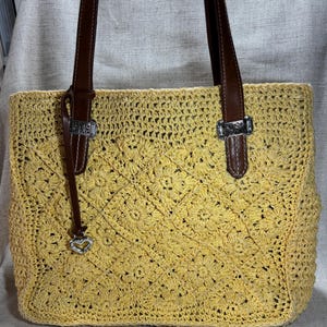 May include: Yellow crocheted tote bag with brown leather handles and silver-tone hardware. The bag features a floral pattern and a small heart charm. The handles are attached with silver buckles.