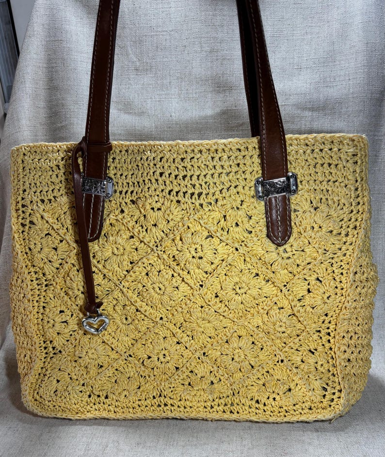 May include: Yellow crocheted tote bag with brown leather handles and silver-tone hardware. The bag features a floral pattern and a small heart charm. The handles are attached with silver buckles.