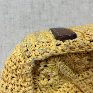 May include: Close-up of a woven yellow bag with a brown leather strap. The bag has a textured, open weave design. The strap is rectangular and stitched to the bag.