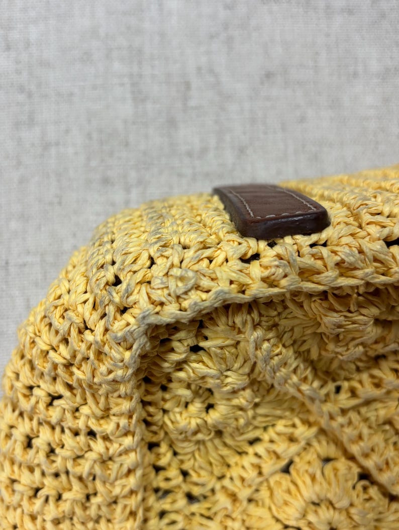 May include: Close-up of a woven yellow bag with a brown leather strap. The bag has a textured, open weave design. The strap is rectangular and stitched to the bag.