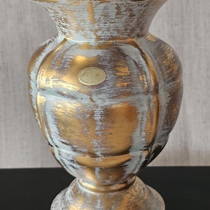May include: A decorative vase with a gold and white distressed finish. The vase has a bulbous body, a pedestal base, and a flared rim. The gold paint is visible through the white paint, creating a textured effect.