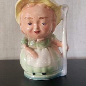May include: A vintage ceramic figurine of a person wearing a yellow hat and a green dress with a white apron. The figurine has a detailed face with blue eyes and is holding a white staff. The base is light blue.