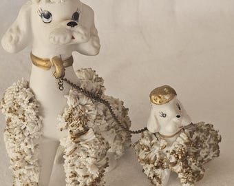 May include: Two white ceramic poodle figurines, one large and one small, connected by a metal chain. The poodles have gold accents on their collars and fur, with black eyes and pink noses. The larger poodle stands upright, while the smaller one is at its side.