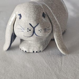 May include: A ceramic rabbit figurine in a relaxed pose. The bunny is primarily white with a crackle glaze effect, and dark blue accents on the ears, eyes, and nose. The figurine has a detailed, textured surface.