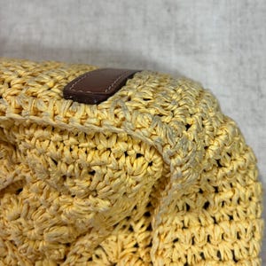 May include: Close-up of a yellow woven bag with a brown leather handle. The bag is made of a thick, textured material with a repeating pattern. The leather handle is rectangular and stitched to the bag.