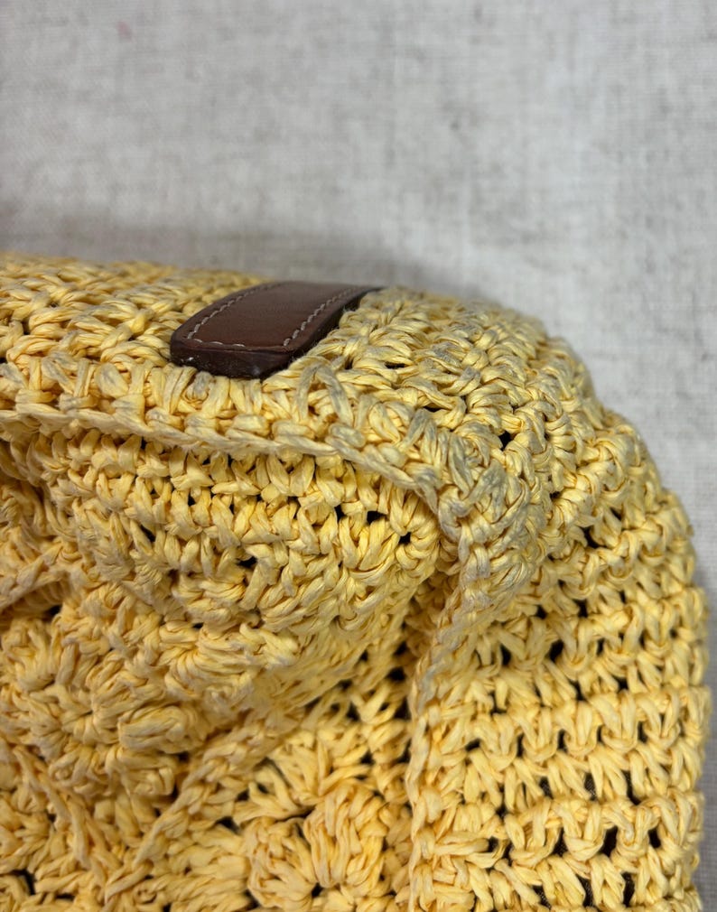 May include: Close-up of a yellow woven bag with a brown leather handle. The bag is made of a thick, textured material with a repeating pattern. The leather handle is rectangular and stitched to the bag.