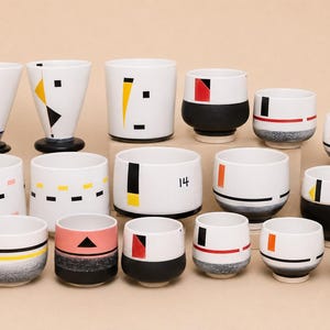 May include: A collection of white ceramic cups and bowls featuring various geometric patterns in black, yellow, red, orange, and pink. Some pieces have solid black bases or gradient textures, while one bowl is clearly marked with the number 14.