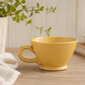 May include: A mustard yellow ceramic mug with a wide, flared rim, a rounded handle, and a pedestal base. The mug features a smooth, solid-colored finish with a slightly tapered shape, providing a stable foundation for holding warm beverages.