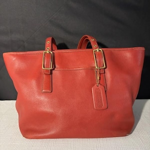 May include: A red leather tote bag with gold-tone hardware. The bag features two top handles, a front pocket, and a leather tag. The bag is a classic style, suitable for everyday use. The bag is made of leather.
