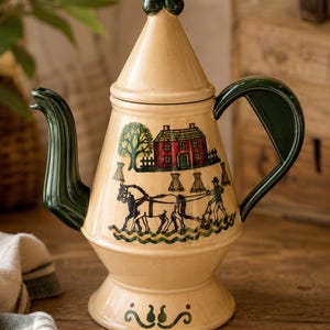 May include: A cream-colored ceramic teapot with a conical lid topped by a dark green finial. The body features a dark green handle and spout, with a painted scene of a red house, a tree, and a person plowing a field with a horse in black and green.