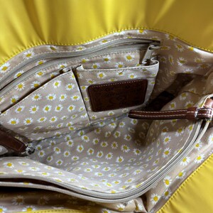 May include: Yellow tote bag with a daisy-patterned interior. The bag has multiple compartments, a brown leather label, and brown leather accents. The interior features a beige fabric with white and yellow daisy flowers.