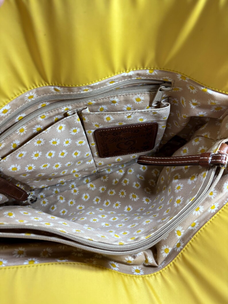 May include: Yellow tote bag with a daisy-patterned interior. The bag has multiple compartments, a brown leather label, and brown leather accents. The interior features a beige fabric with white and yellow daisy flowers.