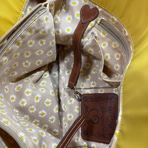 May include: Yellow tote bag with a daisy print interior. The bag features a brown leather strap with a heart-shaped accent and a brown leather tag with the Brighton logo.