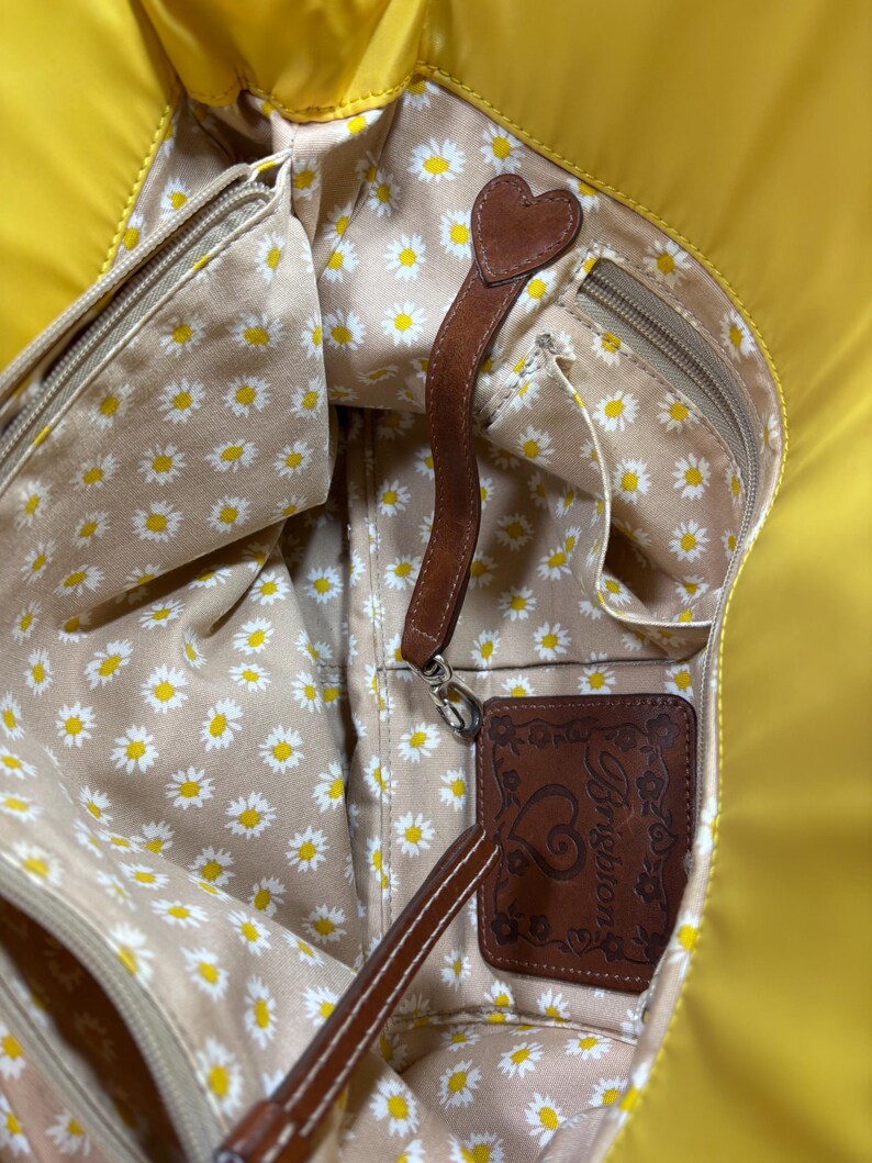 May include: Yellow tote bag with a daisy print interior. The bag features a brown leather strap with a heart-shaped accent and a brown leather tag with the Brighton logo.