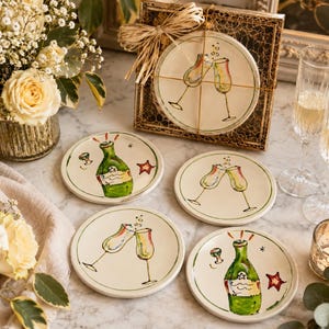 May include: A set of four round ceramic coasters featuring hand-painted illustrations of green champagne bottles and clinking glasses. One coaster is packaged in a square wire frame with a raffia bow, while three are displayed on a marble surface.