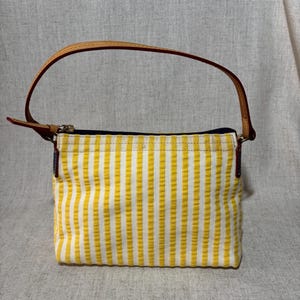 May include: A small, rectangular handbag with vertical yellow and white stripes. It has a brown leather handle and a zipper closure. The bag's design is simple and classic, suitable for everyday use.