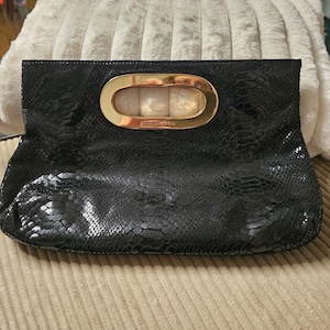 May include: Black clutch with a snakeskin pattern. The clutch has a gold-colored handle with the brand name. The clutch is rectangular and has a fold-over design. The clutch is on a beige surface.