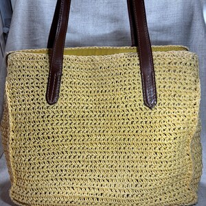 May include: A woven straw tote bag with brown leather handles. The bag is a light yellow color and has a rectangular shape. The handles are attached to the bag with brown stitching.