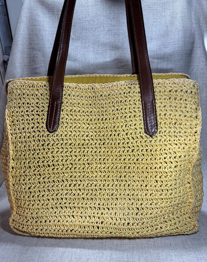 May include: A woven straw tote bag with brown leather handles. The bag is a light yellow color and has a rectangular shape. The handles are attached to the bag with brown stitching.