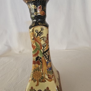 May include: A decorative ceramic candlestick holder with a black, gold, and cream color scheme. The candlestick features floral and geometric patterns, with a flared base and a detailed top. The design includes accents of red, green, and blue.