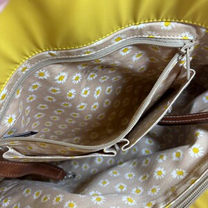 May include: Yellow handbag with a daisy-patterned interior. The bag has multiple pockets and a brown leather strap. The interior lining is beige with a repeating pattern of white and yellow daisies. The exterior is a solid, bright yellow.