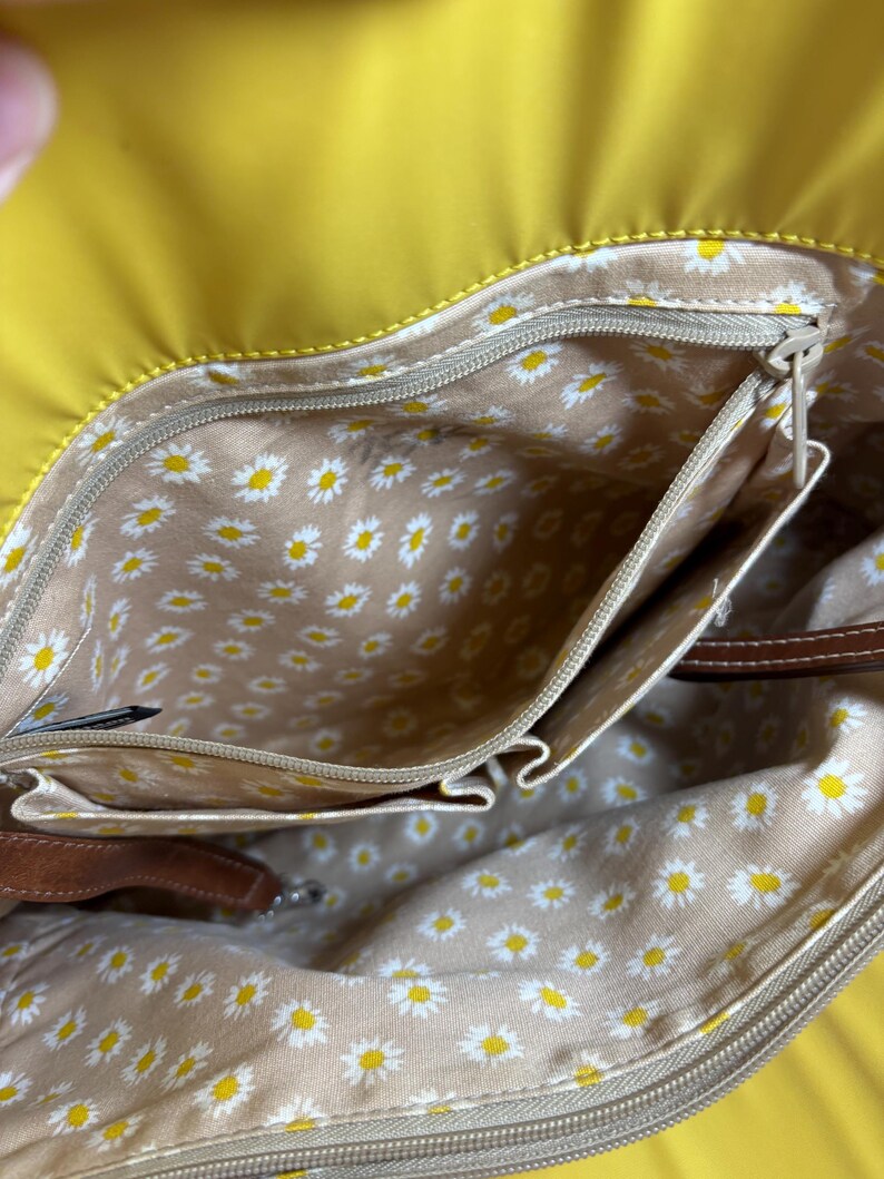 May include: Yellow handbag with a daisy-patterned interior. The bag has multiple pockets and a brown leather strap. The interior lining is beige with a repeating pattern of white and yellow daisies. The exterior is a solid, bright yellow.
