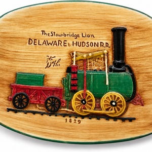 May include: An oval ceramic dish with a wood-grain finish featuring a relief of the Stourbridge Lion steam locomotive. The train is green, red, and yellow with black wheels and a tall chimney. Text reads The Stourbridge Lion, DELAWARE & HUDSON R.R., and 1829.