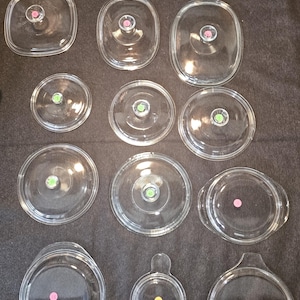 May include: A collection of clear glass cookware lids in various shapes and sizes, including round, square, and oval. Each lid has a knob or handle and a colored sticker. The lids are arranged on a dark surface.