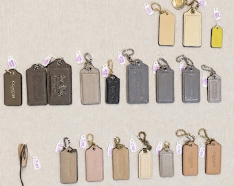 Könnte beinhalten: Eine Sammlung rechteckiger Lederanhänger der Marke Coach in verschiedenen Größen und Farben, darunter Schwarz, Braun, Beige, Grau und Blau. Jeder Anhänger ist mit einer Metallkugelkette und einem kleinen weißen Papieretikett mit Codes versehen.