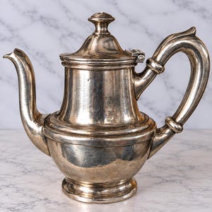 May include: A polished silver teapot featuring a rounded base, a tapered cylindrical body, and a curved spout. The teapot includes a matching lid with a small knob handle and a curved side handle, all finished in a reflective metallic silver.