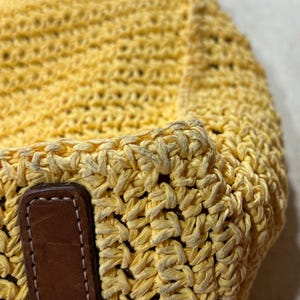 May include: Close-up of a yellow woven tote bag with brown leather handles. The bag is made of a thick, textured material, and the leather handles are stitched to the bag. The bag is a light yellow color.