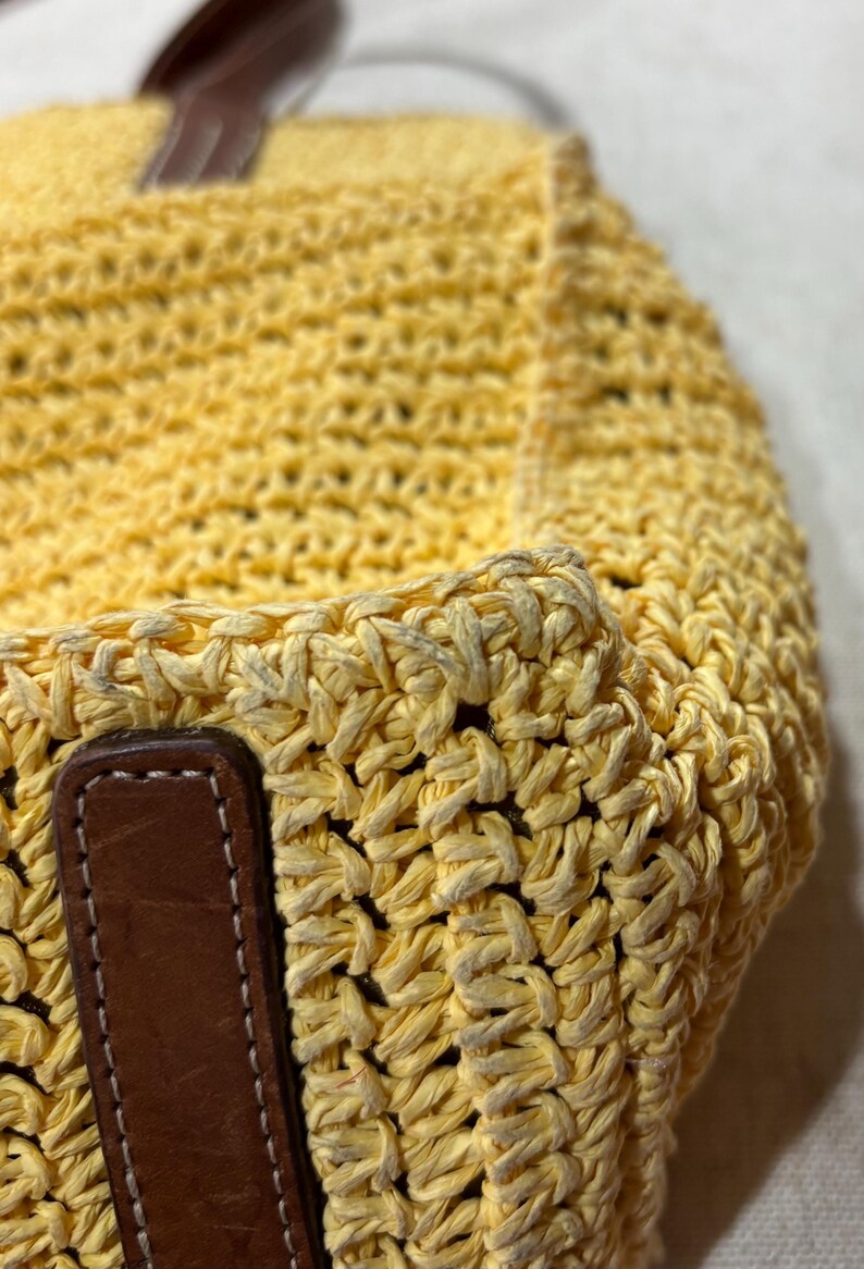 May include: Close-up of a yellow woven tote bag with brown leather handles. The bag is made of a thick, textured material, and the leather handles are stitched to the bag. The bag is a light yellow color.