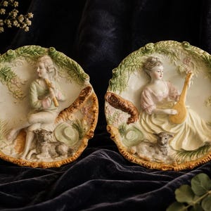 May include: Two circular porcelain wall plates featuring raised relief scenes of figures in a pastoral setting. One plate depicts a person playing a flute, the other a person playing a lute, both accompanied by a small sheep and floral details.