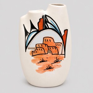 May include: A white ceramic vase with two openings at the top. The front features a hand-painted design of orange adobe-style buildings, black desert vegetation, and a large black and blue archway against a light background, creating a desert scene.