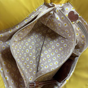 May include: Bright yellow tote bag with multiple interior pockets. The interior lining features a daisy pattern on a tan background. A brown leather heart-shaped accent is visible. The bag has a woven trim.