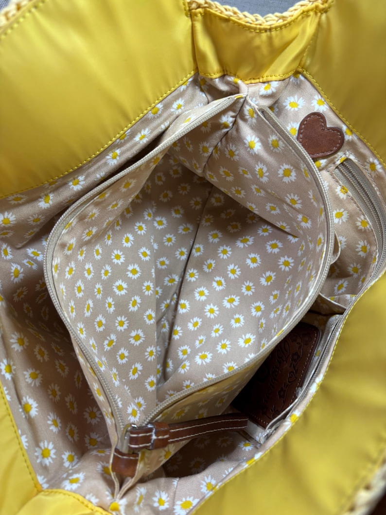 May include: Bright yellow tote bag with multiple interior pockets. The interior lining features a daisy pattern on a tan background. A brown leather heart-shaped accent is visible. The bag has a woven trim.