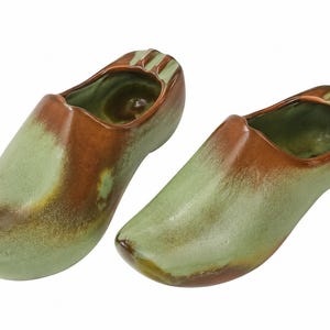 May include: A pair of ceramic clogs featuring a glazed finish with blended sage green and warm brown tones. The shoes have a smooth, rounded toe design and an open top, showcasing a glossy, reflective surface throughout the entire exterior.