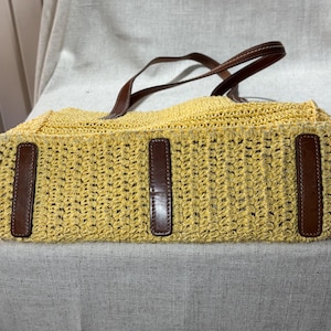 May include: A yellow woven tote bag with brown leather accents. The bag features a rectangular shape with a woven pattern and brown leather handles. The bag has three brown leather accents on the front.