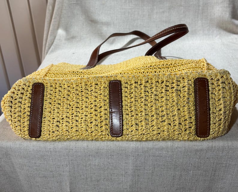 May include: A yellow woven tote bag with brown leather accents. The bag features a rectangular shape with a woven pattern and brown leather handles. The bag has three brown leather accents on the front.