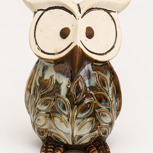 May include: A ceramic owl figurine featuring a cream-colored face with large circular eyes and a dark brown beak. The body is finished in a glossy brown and light blue glaze, decorated with a repeating embossed leaf pattern across the front.