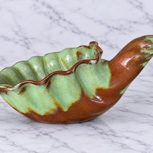 May include: A ceramic bowl shaped like a shell with a ruffled edge. The interior features a light mint green glaze, while the exterior and the elongated handle are finished in a glossy, deep brown glaze with visible color blending.