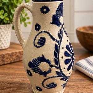 May include: A beige ceramic pitcher featuring a tall, slender neck and a curved handle. The exterior is decorated with deep blue hand-painted floral motifs, swirling vine patterns, and small circular accents, all set against a smooth, light-colored clay body.
